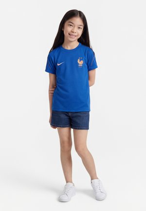 Fiatal lány áll, kék Franciaország focimezben, farmer rövidnadrágban és fehér tornacipőben, mosolyogva, kezét a hát mögött tartva.