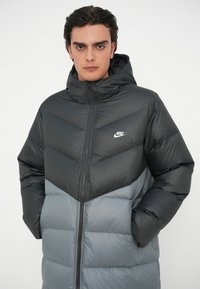 Crna i siva jakna s kapuljačom i jastučićima s ševron uzorkom, sa patentnim zatvaračem sprijeda i Nike logom na prsima. Mekana, quilted tekstura.