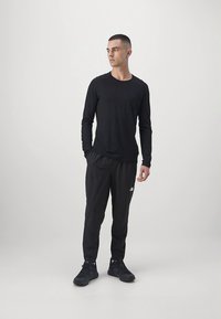 Camisola preta de manga comprida com decote redondo, combinada com calças de jogging pretas com um logótipo. O modelo usa sapatilhas desportivas pretas.