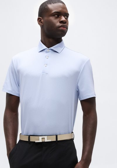 Νέος άνδρας με ανοιχτό μπλε πικέ πόλο πουκάμισο, μπεζ ζώνη με γεωμετρική αγκράφα και μαύρο παντελόνι, κοιτάζοντας προς τα πλάγια, μπροστά σε απλό φόντο.