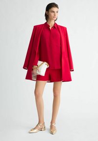 Blazer rosso, camicia abbinata e pantaloni corti, con una borsa bianca dotata di una catena dorata. Scarpe piatte oro con fiocchi completano l'outfit.