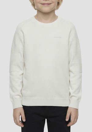 Bambino che indossa un maglione bianco a coste a manica lunga con maniche strutturate e pantaloni neri, in piedi davanti a uno sfondo semplice.