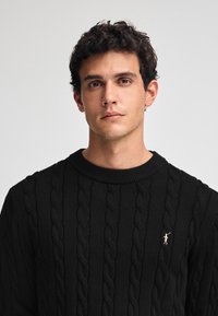 Svart kabelstickad tröja med rund halsringning, med en subtil broderad logotyp på bröstet. Tyget har en texturerad yta.