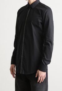 Camisa negra de manga larga con cuello estándar, que presenta un detalle de cremallera plateada a lo largo del tapeta delantera y un tejido liso y texturizado.