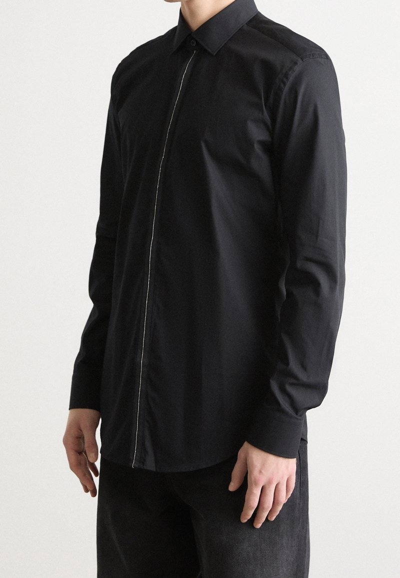 Camisa negra de manga larga con cuello estándar, que presenta un detalle de cremallera plateada a lo largo del tapeta delantera y un tejido liso y texturizado.