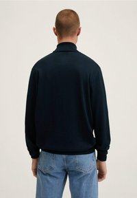 Tummansininen pitkähihainen polo paita, jossa on korkea kaulus, rento leikkaus ja ribatut hihansuut, yhdistettynä vaaleansinisiin farkkuihin.