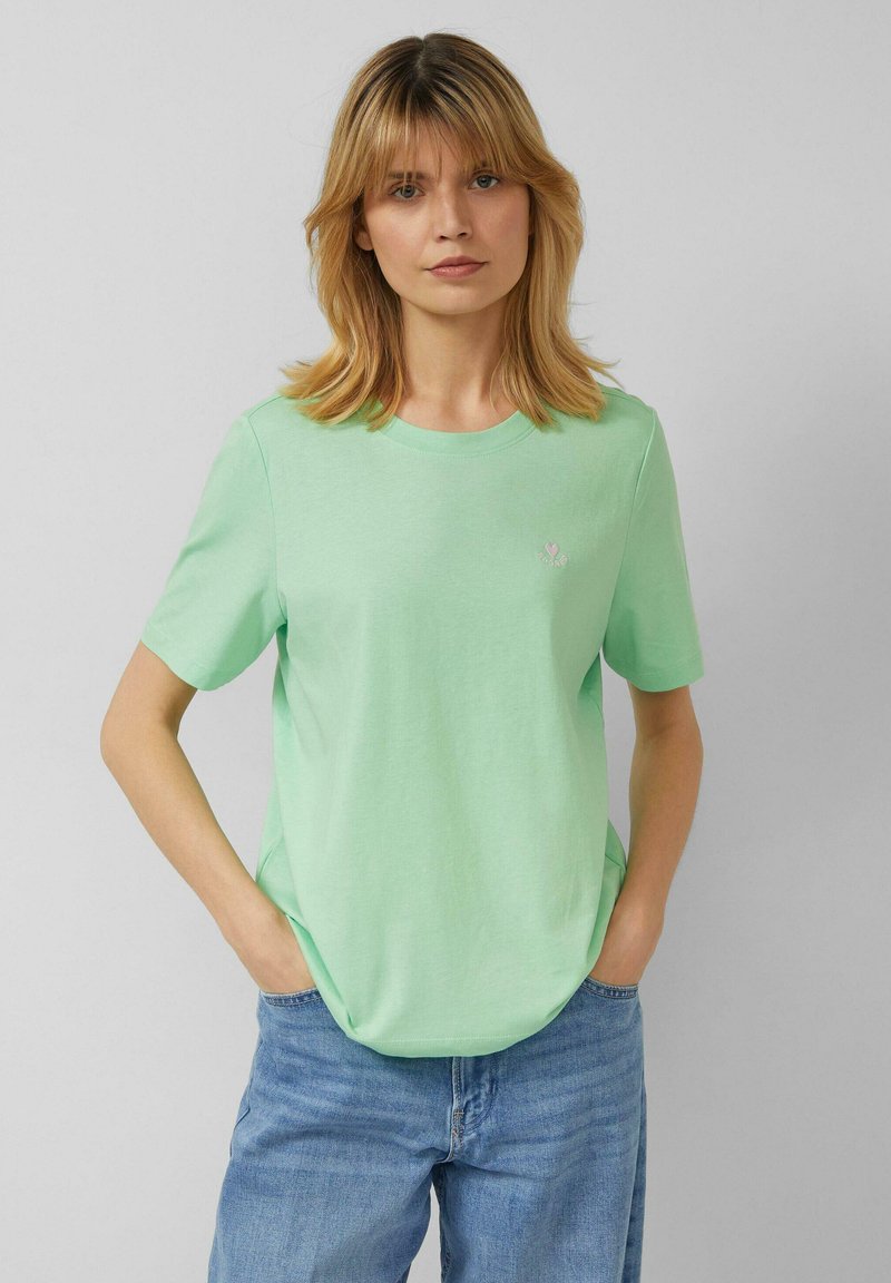 Mujer rubia con camiseta de manga corta verde claro con diseño de corazón pequeño, manos en los bolsillos y vaqueros azules relajados contra un fondo liso.