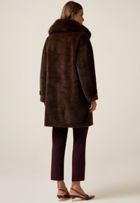 Kobieta w ciemnobrązowym futerku z tworzywa sztucznego, bordowych krótkich spodniach i bordowych szpilkach z paseczkiem za piętą, stojąca na jednolitym tle.