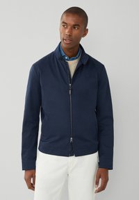 Mørkeblå jakke med lynlås foran, ståkrave, to sidelommer og en glat overflade, båret over en lys sweater og en skjorte med krave.
