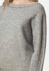 Pullover grigio chiaro a maglia con scollatura ampia, decorato con borchie metalliche lungo le spalle e lungo le maniche. Texture morbida.