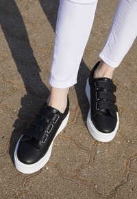 Svarta lädersneakers med tre breda remmar, vit texturerad sula, rund tå och slät yta som vilar på en strukturerad yta.