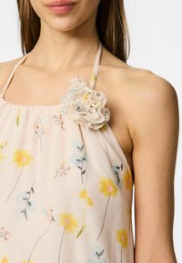 Blommig halterneck-top i ljusrosa tyg med gula och blå blommönster, med en texturerad blomaccent på vänster axel.