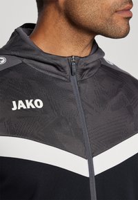 Sort og grå lynlåsjakke med tekstureret overdel, hvid stribe detalje og mærket "JAKO" på brystet.