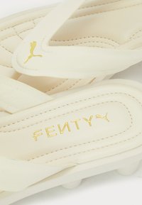 Cream-colored slide sandals made of soft material, featuring a quilted design, gold "FENTY" logo, and Puma's signature logo on the strap.