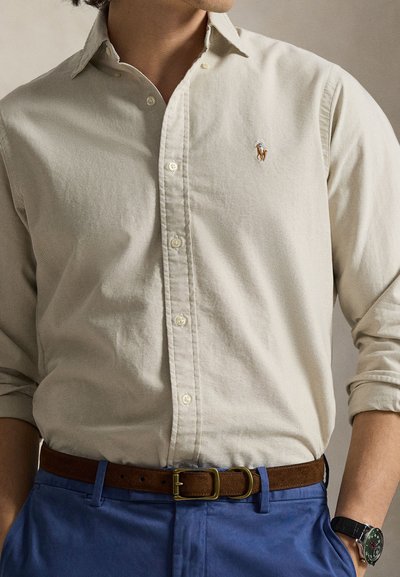Μπεζ πουκάμισο με κουμπιά και κλασικό γιακά, μικρό κεντητό λογότυπο, συνδυασμένο με μπλε παντελόνι και καφέ ζώνη. Ρολόι ορατό στον καρπό.