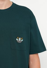 Temno zelena kratkosleeva majica z žepom na prsih in barvnim vezenim logotipom, ki vsebuje insignijo s cvetjem in črtami.