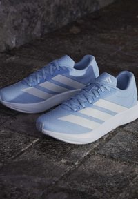 Zapatillas deportivas de color azul claro con una parte superior de malla texturizada, con tres rayas blancas a ambos lados y una suela de goma blanca.