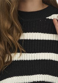 Longs cheveux bruns sur un pull en maille noir et blanc à rayures horizontales, avec une main ajustant le col sur le côté droit.