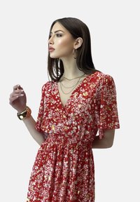 Röd blommig klänning med korta ärmar, med djup V-ringning och smockad midja. Accentuerad med vita och gyllene blommor. Gyllene hoopörhängen och armbandsur.