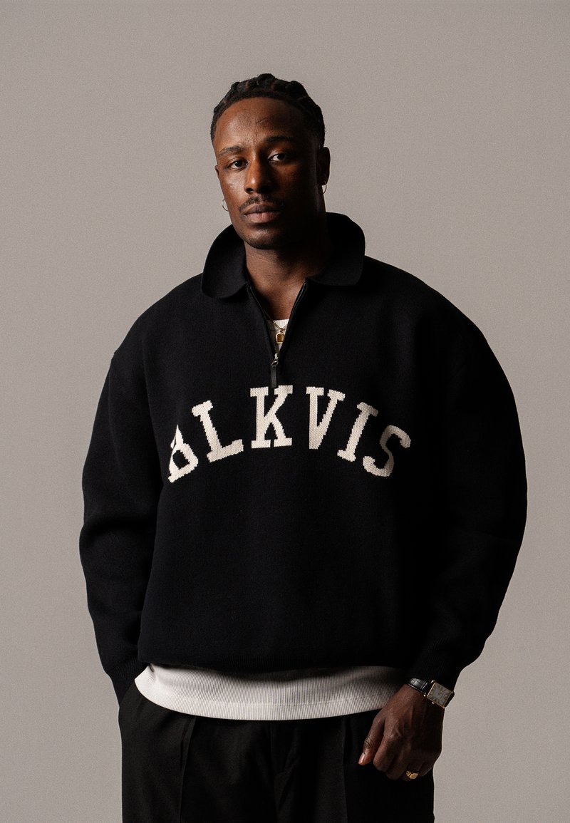 Schwarzer Pullover mit hohem Kragen und Reißverschluss auf der Vorderseite, mit weißen "ALKVIS"-Buchstaben auf der Brust und geripptem Saum.