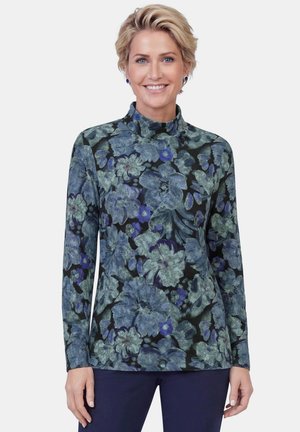 Woman with short blonde hair smiles wearing a long-sleeve floral print turtleneck top in blue and green shades and dark pants.