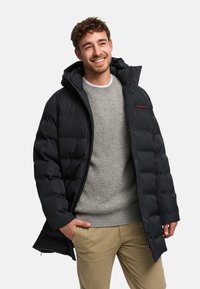 Mustavärinen toppatakki, jossa vetoketjullinen etuosa ja huppu, quiltattu kuviointi sekä punainen logodetalji. Yhdistetty harmaaseen ribbineulottuun neulepaitaan.