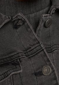 Chaqueta de mezclilla gris oscuro con cierre de botones, cuello acanalado y costuras visibles. Incluye un detalle de costura rota en un hombro.