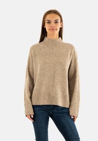 Suéter beige de cuello alto hecho de un suave tejido de punto, con un corte holgado y un dobladillo acanalado, combinado con jeans azul oscuro.