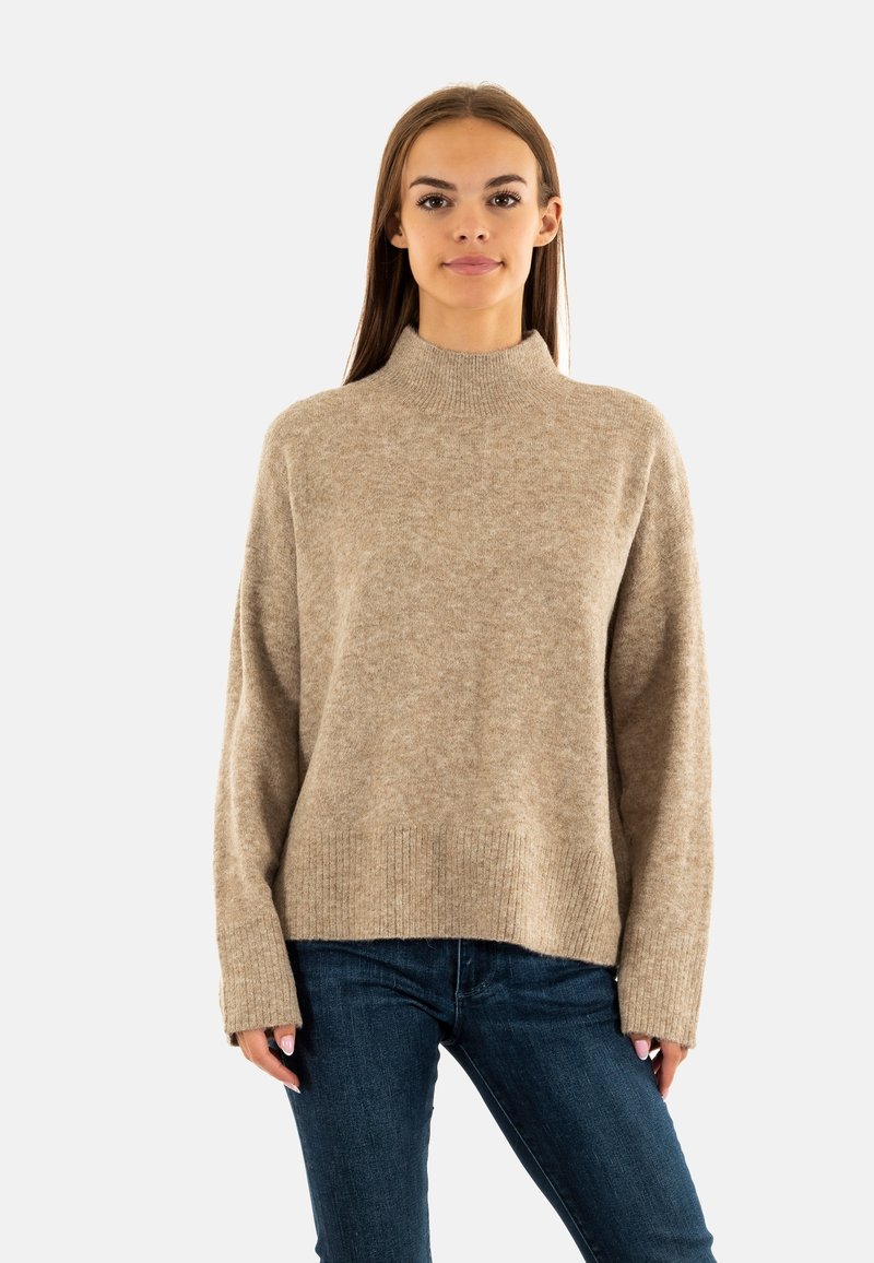 Suéter beige de cuello alto hecho de un suave tejido de punto, con un corte holgado y un dobladillo acanalado, combinado con jeans azul oscuro.