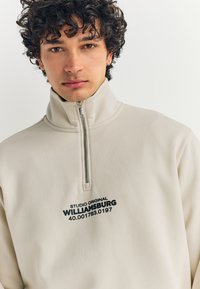 Hellbeige Fleecejacke mit Reißverschluss und hohem Kragen, mit schwarzem Aufdruck: "STUDIO ORIGINAL WILLIAMSBURG 40.001783.0197." Glatte Textur.