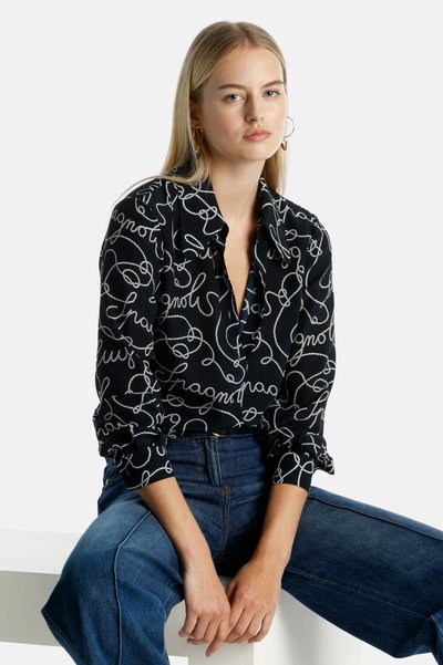 Blusa nera con testo bianco in corsivo. Presenta un colletto e polsini con bottoni. Indossata con jeans blu a vita alta.