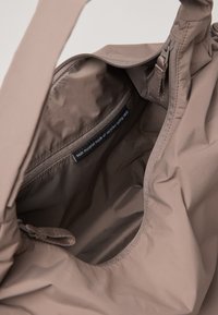 Bruine tas van gerecycled stof met een ritssluiting, een soepele textuur en een binnenzaklabel dat aangeeft dat het gemaakt is van gerecyclede visnetten.