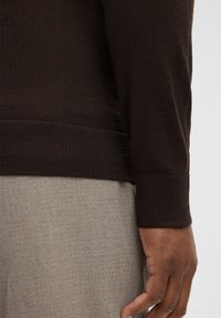 Rechte Hand einer Person, die einen dunkelbraunen Langarmpullover und hellgraue, strukturierte Hose trägt, vor einem einfarbigen Hintergrund.
