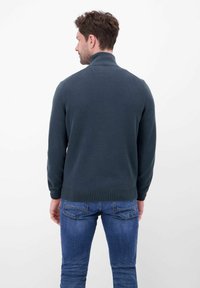 Donkergroene gebreide trui met een hoge kraag en een textuurpatroon, gecombineerd met een blauwe spijkerbroek. De trui heeft geribbelde manchetten en zoom.