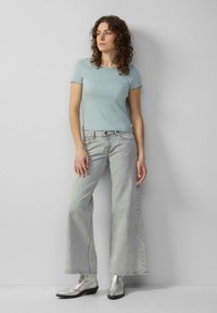 Frau mit lockigem Haar, die ein hellblaues kurzärmliges Oberteil, graue weit geschnittene Jeans und silberne Stiefeletten trägt, steht vor einer hellgrauen Wand.