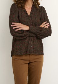 Woman with crossed arms wearing a long-sleeve brown blouse with red floral patterns and brown pants against a plain background.