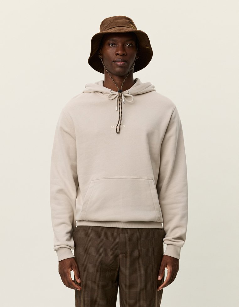 Beige kapucnis pulóver zsinórral, elülső zsebbel és bordázott mandzsettával párosítva barna kockás nadrággal és barna kalappal.