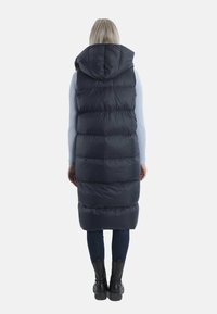 Marineblauer Puffer-Weste mit Kapuze, ausgestattet mit einem langen Design, gesteppten Texturen und einem gerafften Saum, getragen über einem hellen Pullover und Jeans.