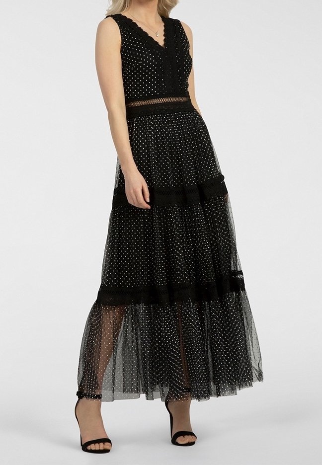 Femme portant une robe noire à pois sans manches avec une jupe superposée transparente et des sandales noires à talons hauts, debout sur un fond uni.