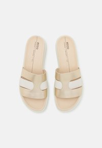 Tan and white slide sandals with a smooth leather upper, featuring cut-out accents and a padded insole. Sole has slight elevation.
