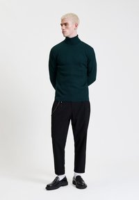 Maglione a collo alto in maglia verde scuro con motivo testurizzato, abbinato a pantaloni neri slim-fit e scarpe nere, in piedi su uno sfondo chiaro.