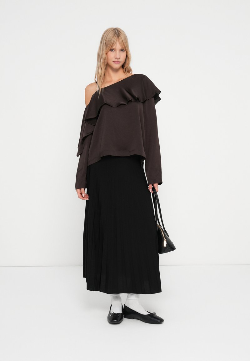 Haut brun à épaules dénudées avec des volants, associé à une jupe plissée noire. Les accessoires comprennent un sac à main noir et des ballerines noires avec des chaussettes blanches.