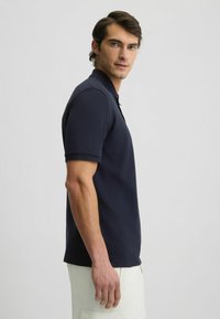 Polo shirt navy in un tessuto morbido e elastico con collo classico e maniche corte, abbinato a pantaloni beige, vista di lato.
