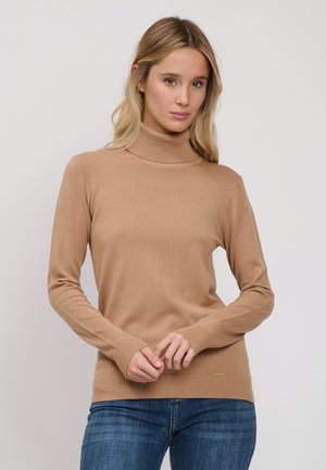 Vrouw met blond haar die een beige coltrui en blauwe spijkerbroek draagt, staand tegen een effen lichte achtergrond.