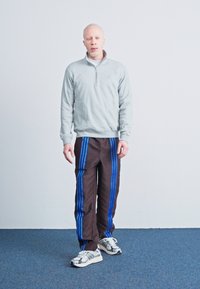 Ανοιχτό γκρίζα φλις ζακέτα με φερμουάρ συνδυασμένη με καφέ φόρμες που διαθέτουν μπλε ρίγες. Λευκά αθλητικά παπούτσια ολοκληρώνουν την εμφάνιση.