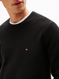 Pull en tricot noir avec une finition texturée, doté d'un col rond blanc et d'un petit logo brodé rouge, blanc et bleu sur la poitrine.