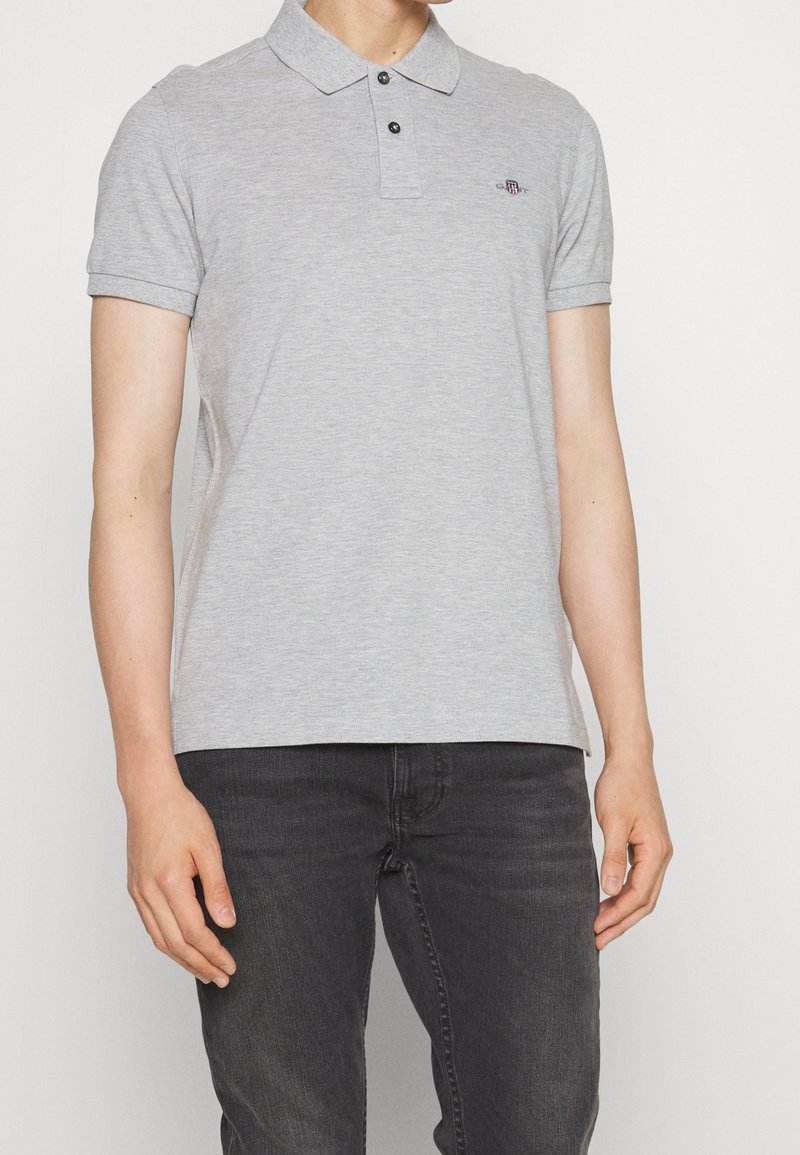 Homme portant un polo gris clair à manches courtes avec deux boutons et un jean gris foncé, debout devant un fond uni.