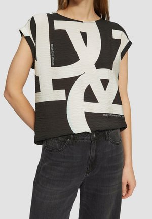 Kobieta ubrana w czarno-biały top z krótkim rękawem o abstrakcyjnym wzorze z napisem „more love stories” oraz ciemnoszare dżinsy.