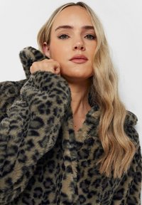 Leopardmönstrad fuskpälsjacka i jordiga toner, med huva, knäppning och mjuk textur. Designen inkluderar svarta och bruna fläckar.
