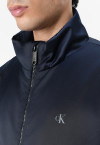Marine waterdichte jas met hoge kraag, rits vooraan en subtiele "CK" logo. Gladde textuur en strak ontwerp voor casual wear.